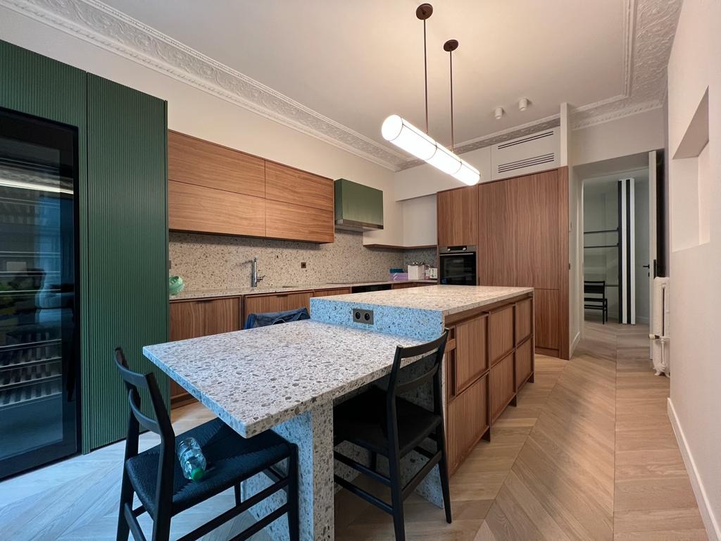 Nowoczesna kuchnia z wyspą i blatem w lastryko, drewniane szafki wiszące, zielona zabudowa lodówki i dekoracyjny sufit. Parkiet w jodełkę na podłodze.