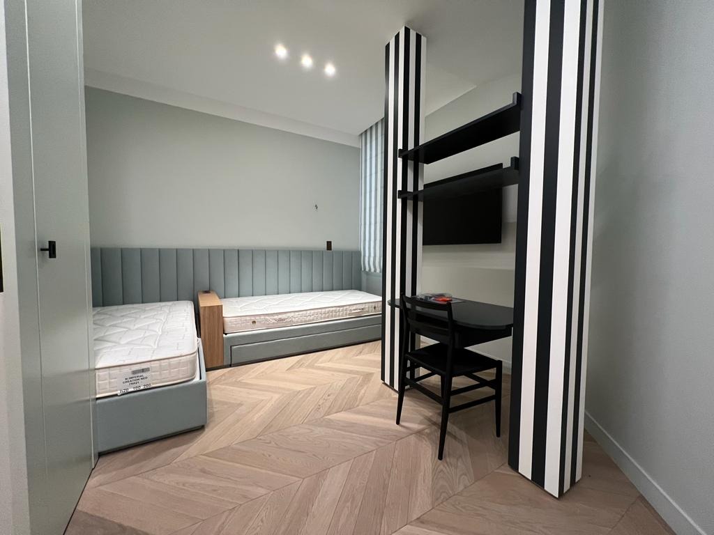 Pokój młodzieżowy z dwoma łóżkami z tapicerowanym wezgłowiem, biurkiem i półkami oddzielonymi czarno-białymi kolumnami. Parkiet w jodełkę na podłodze.