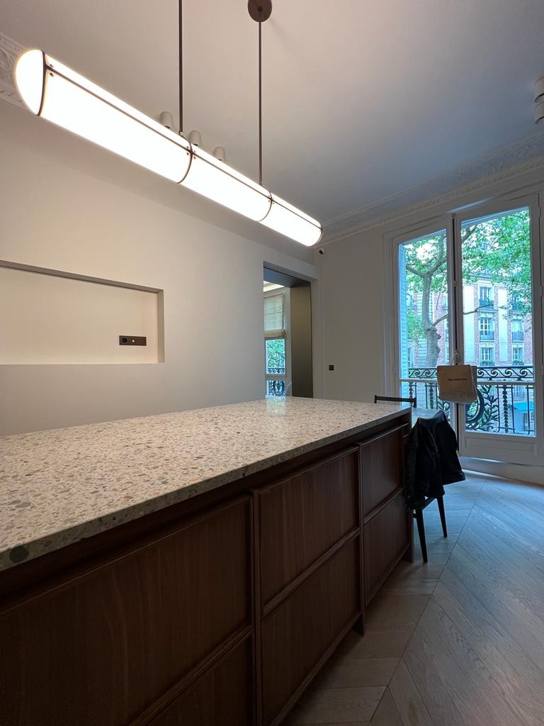 Nowoczesna wyspa kuchenna z blatem lastryko i drewnianymi frontami, oświetlona podłużną lampą. Widok na balkon przez okno.