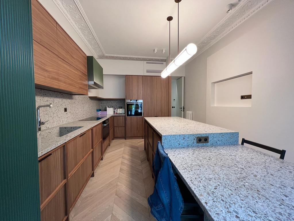 Nowoczesna kuchnia z drewnianymi frontami, blatem w terrazzo i wyspą. Uwagę przykuwa długa lampa sufitowa i dekoracyjny gzyms. Widoczny fragment zielonej ściany.