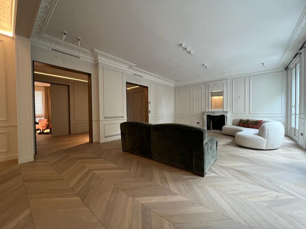 Elegancki salon z parkietem w jodełkę, zieloną sofą i białym narożnikiem. Klasyczne sztukaterie i wysokie listwy przypodłogowe dodają wnętrzu szyku. Widoczne drzwi przesuwne.