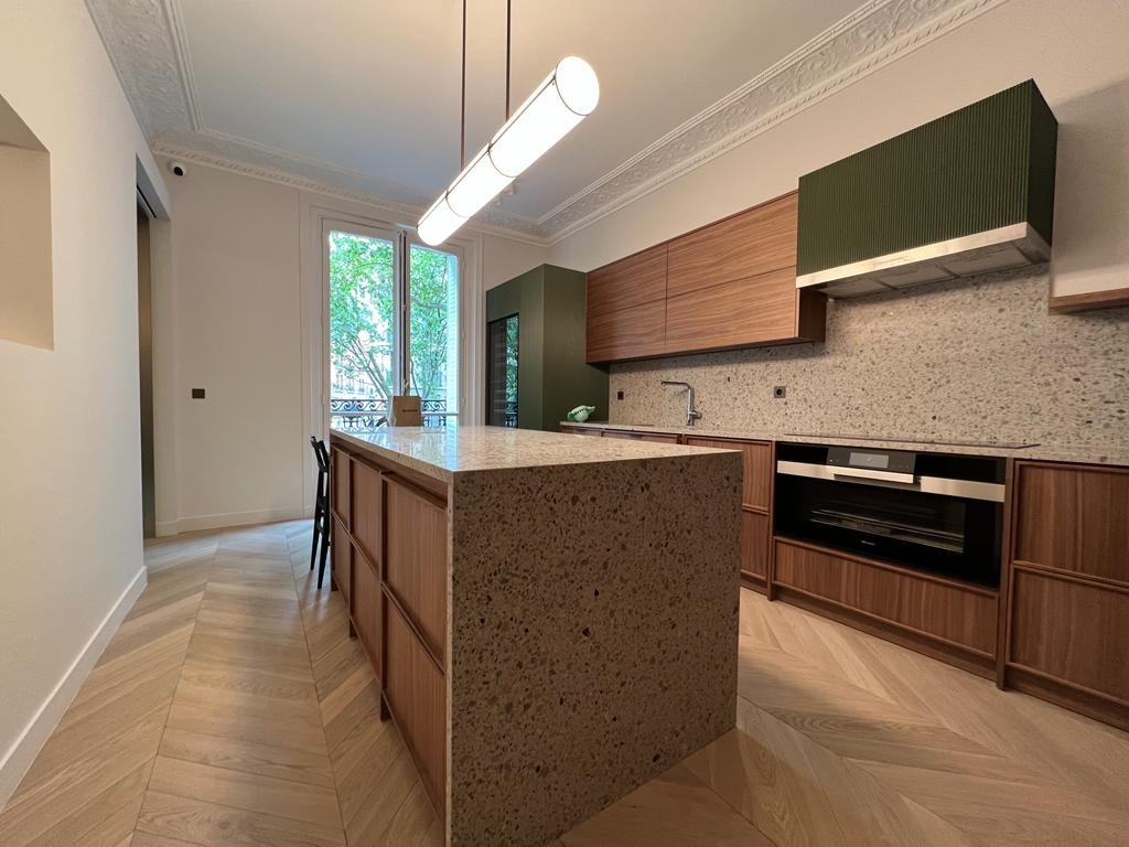 Nowoczesna kuchnia z wyspą o blacie lastryko, drewniane szafki, zielony okap i parkiet w jodełkę. Długi, tubowy żyrandol. Widok na zieleń za oknem.