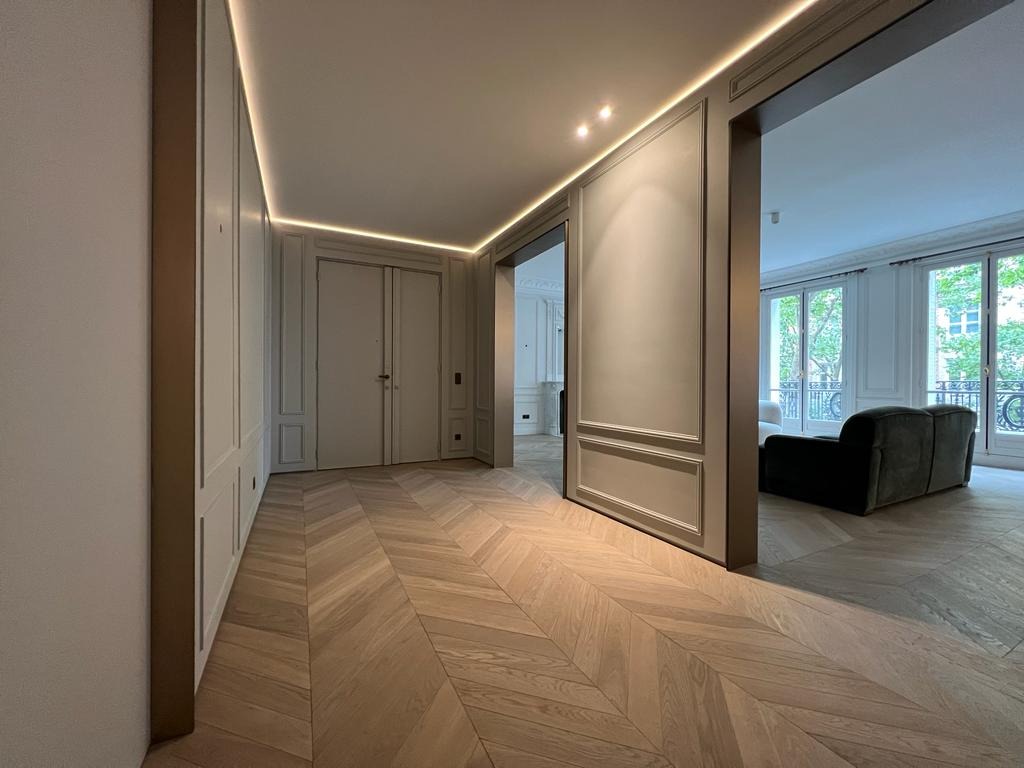 Eleganckie wnętrze z parkietem w jodełkę, listwami ściennymi i subtelnym oświetleniem LED. Widok na salon z sofą i oknami balkonowymi.