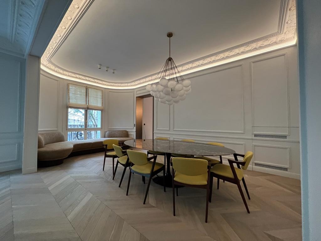 Elegancka jadalnia z owalnym stołem, żółtymi krzesłami i parkietem w jodełkę. W tle sofa przy oknie i dekoracyjne listwy sufitowe z oświetleniem.