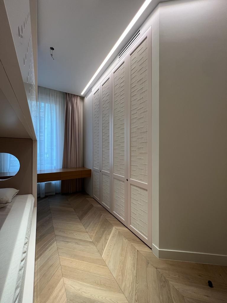 Biała szafa wnękowa z dekoracyjnymi drzwiami harmonijkowymi, oświetlenie LED podsufitowe, parkiet w jodełkę, biurko i łóżko piętrowe w tle. Nowoczesny design.