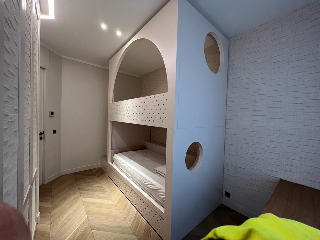 Nowoczesne łóżko piętrowe na wymiar w pokoju dziecięcym, z okrągłymi otworami i subtelnym oświetleniem, na tle wzorzystej tapety i parkietu w jodełkę.