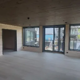 Wnętrze domu szkieletowego z drewnianą boazerią, widok na zewnątrz przez duże okna i drzwi balkonowe, sufit wykończony drewnem.