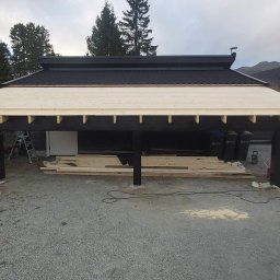 Nord-House - Budowa zadaszenia na czterech czarnych słupach, widoczne drewniane elementy konstrukcyjne dachu, w tle góry, pracownik w odblaskowej kamizelce używa piły elektrycznej do cięcia drewna.