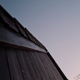 Nord-House - Niski kąt ujęcia na fragment drewnianego domu z ciemnymi deskami elewacyjnymi i widocznym fragmentem dachu, na tle błękitnego nieba z delikatnym różowym zabarwieniem i gałęziami drzew bez liści.