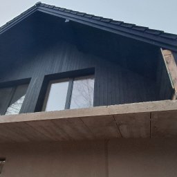 Nord-House - Surowy stan domu szkieletowego z widoczną konstrukcją betonową balkonu i czarnym drewnianym wykończeniem elewacji oraz dachu.