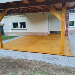 Nord-House - Świeżo wykończony taras drewniany z zadaszeniem, otoczony szarym żwirem, przylegający do budynku z białymi roletami.