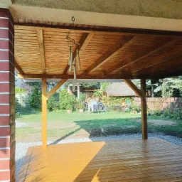 Zadaszony taras z drewnianą podłogą i widokiem na zadbany ogród z elementami małej architektury, w tle widoczny domek z trzcinowym dachem.