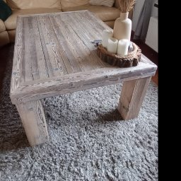 Nord-House - Stolik kawowy z surowego, bielonego drewna, na grubych nogach, ustawiony na puszystym, szarym dywanie. Na blacie dekoracja z suszonych traw w wazonie i świec na drewnianej podstawce. W tle beżowa...
