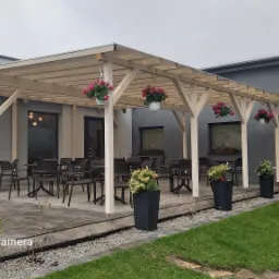 Zadaszony taras przy budynku z szarego tynku, wyposażony w stoliki i krzesła, ozdobiony wiszącymi i stojącymi donicami z kwiatami.