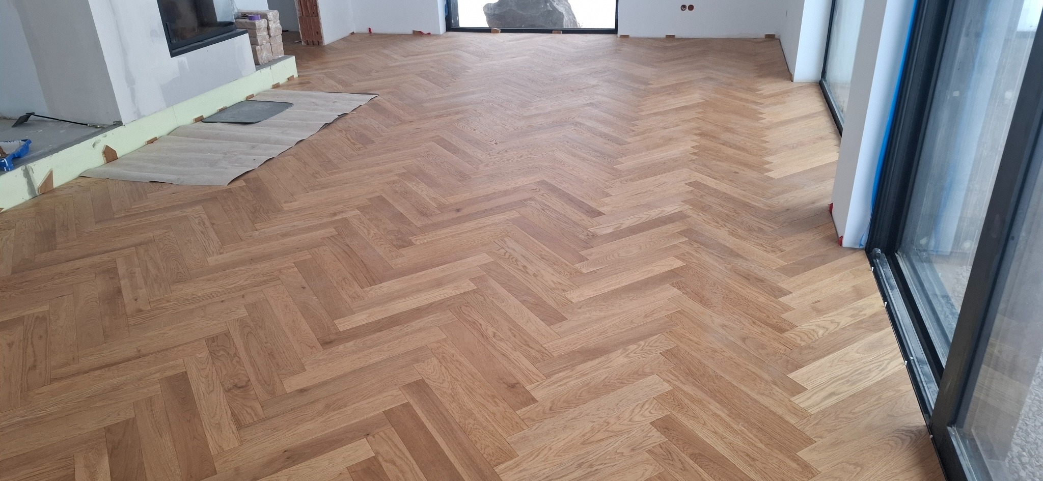 Jasny parkiet ułożony we wzór jodełki francuskiej, częściowo zabezpieczony kartonem przy kominku, widoczne przygotowania do dalszych prac wykończeniowych.