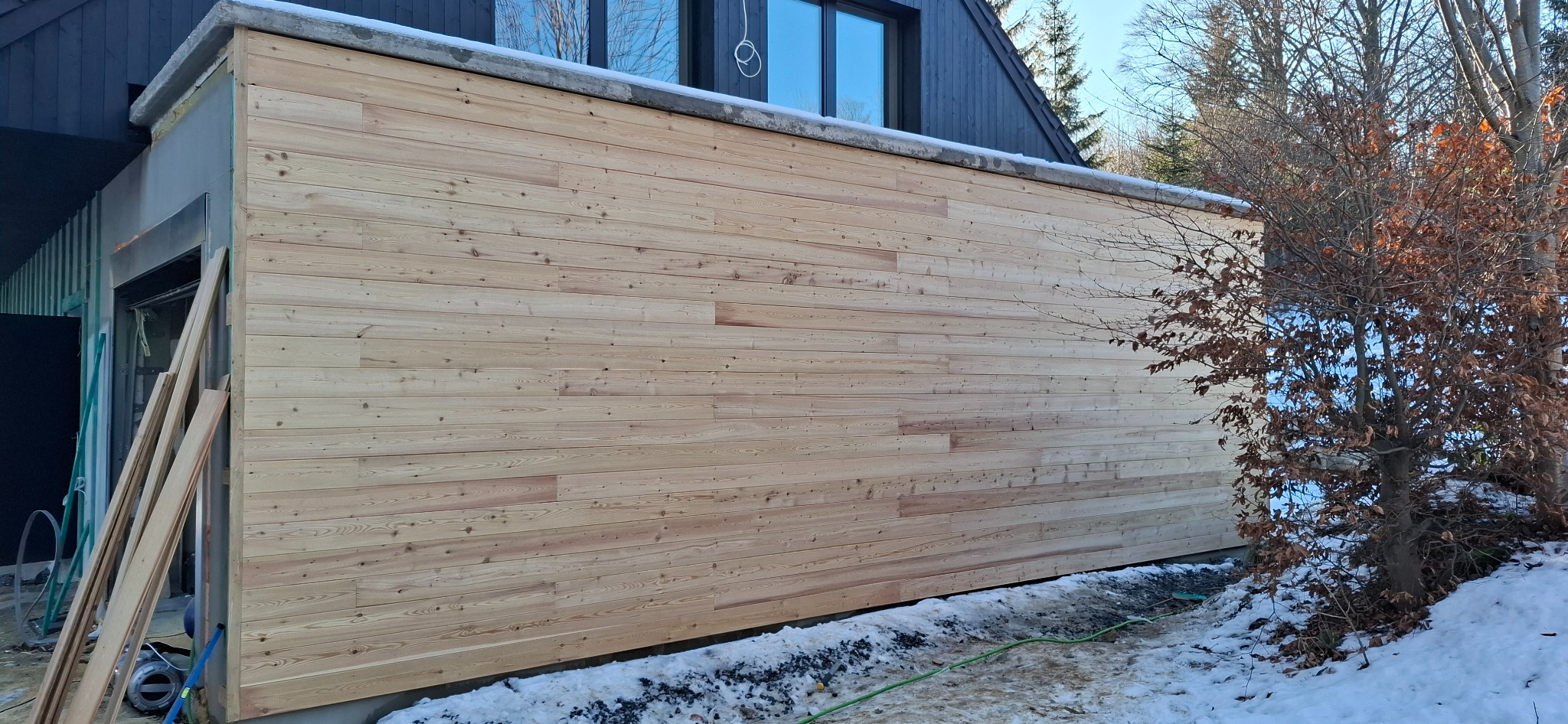 Ściana budynku w trakcie obkładania jasnym drewnem, widoczne deski i fragment elewacji z oknami, śnieg na ziemi.