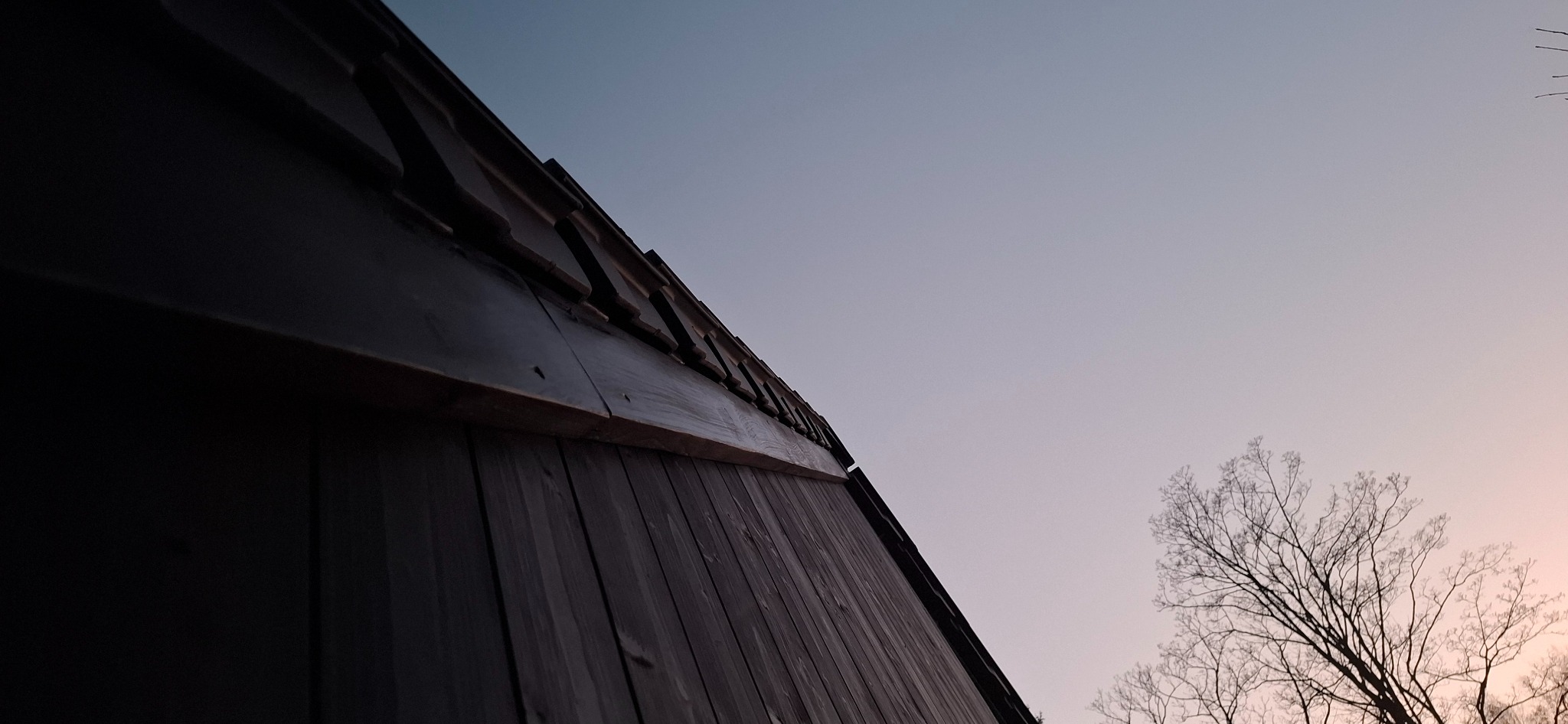 Niski kąt ujęcia na fragment drewnianego domu z ciemnymi deskami elewacyjnymi i widocznym fragmentem dachu, na tle błękitnego nieba z delikatnym różowym zabarwieniem i gałęziami drzew bez liści.