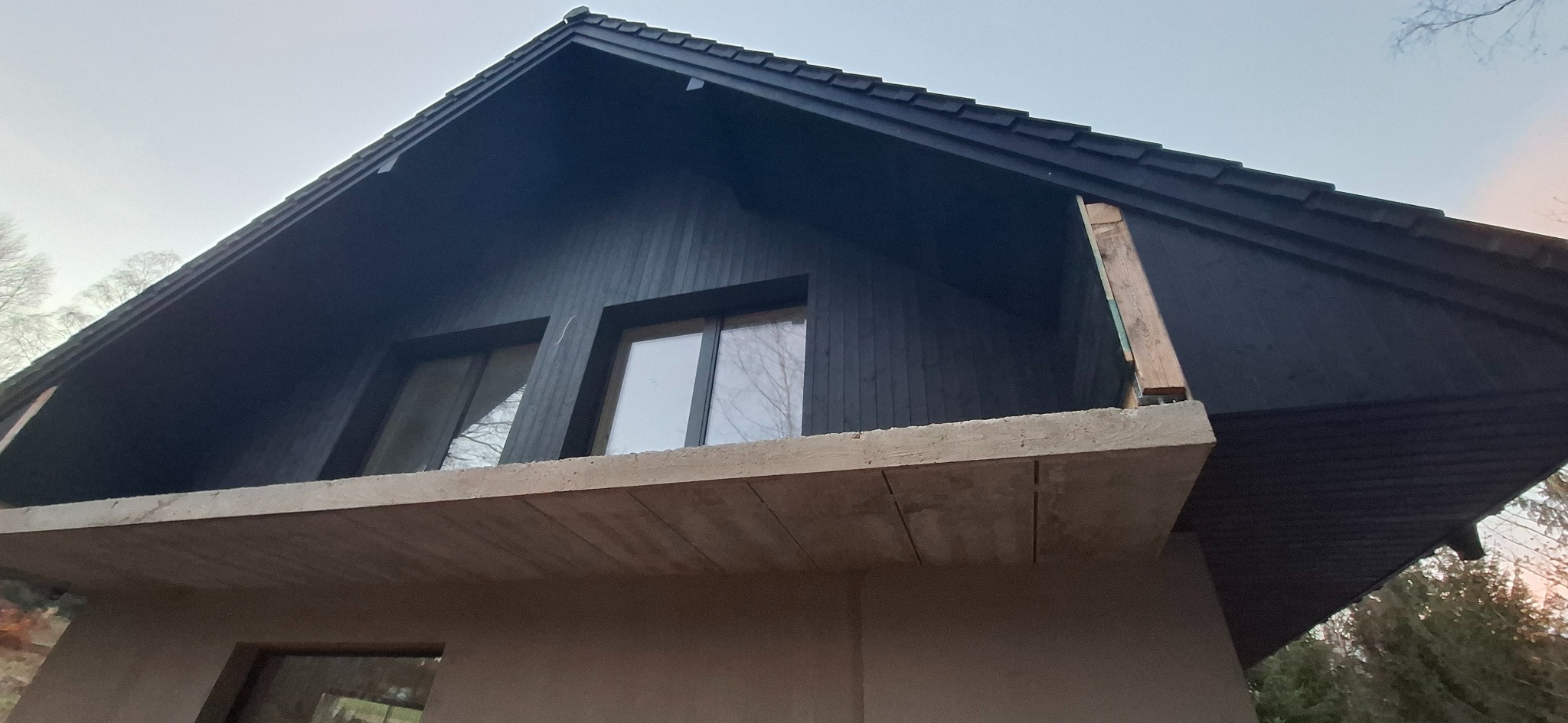 Surowy stan domu szkieletowego z widoczną konstrukcją betonową balkonu i czarnym drewnianym wykończeniem elewacji oraz dachu.