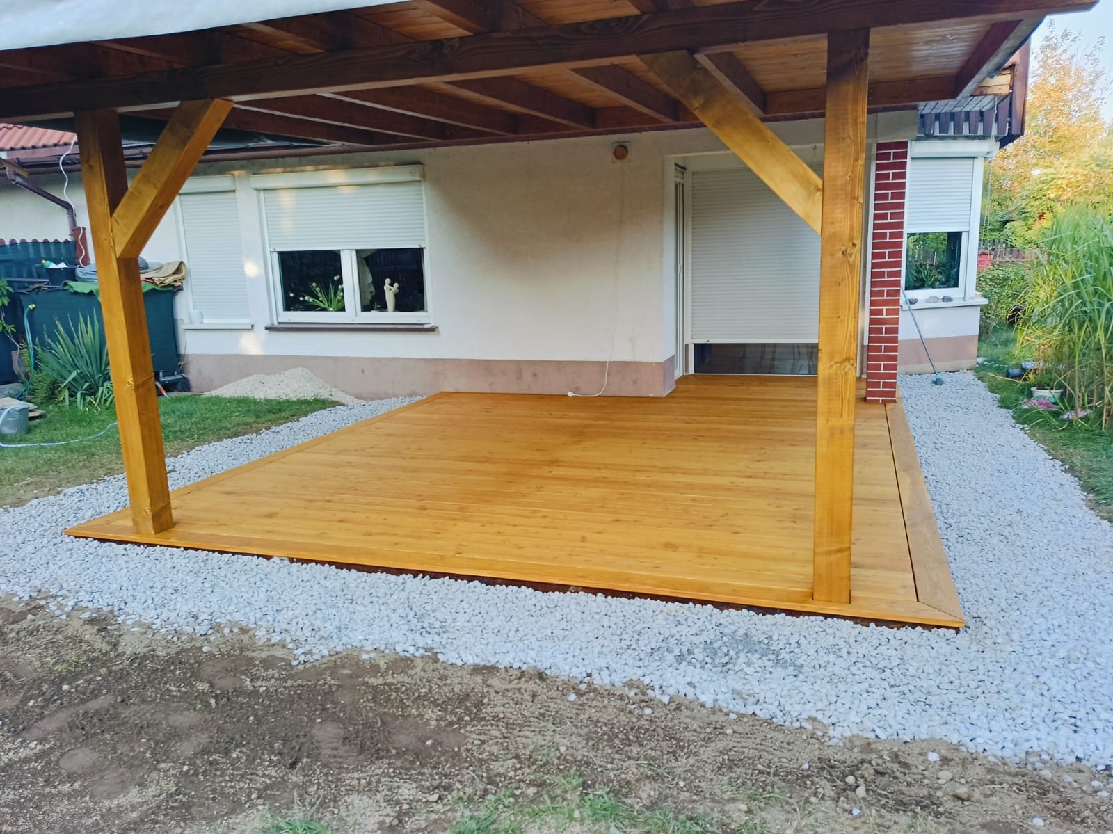 Świeżo wykończony taras drewniany z zadaszeniem, otoczony szarym żwirem, przylegający do budynku z białymi roletami.