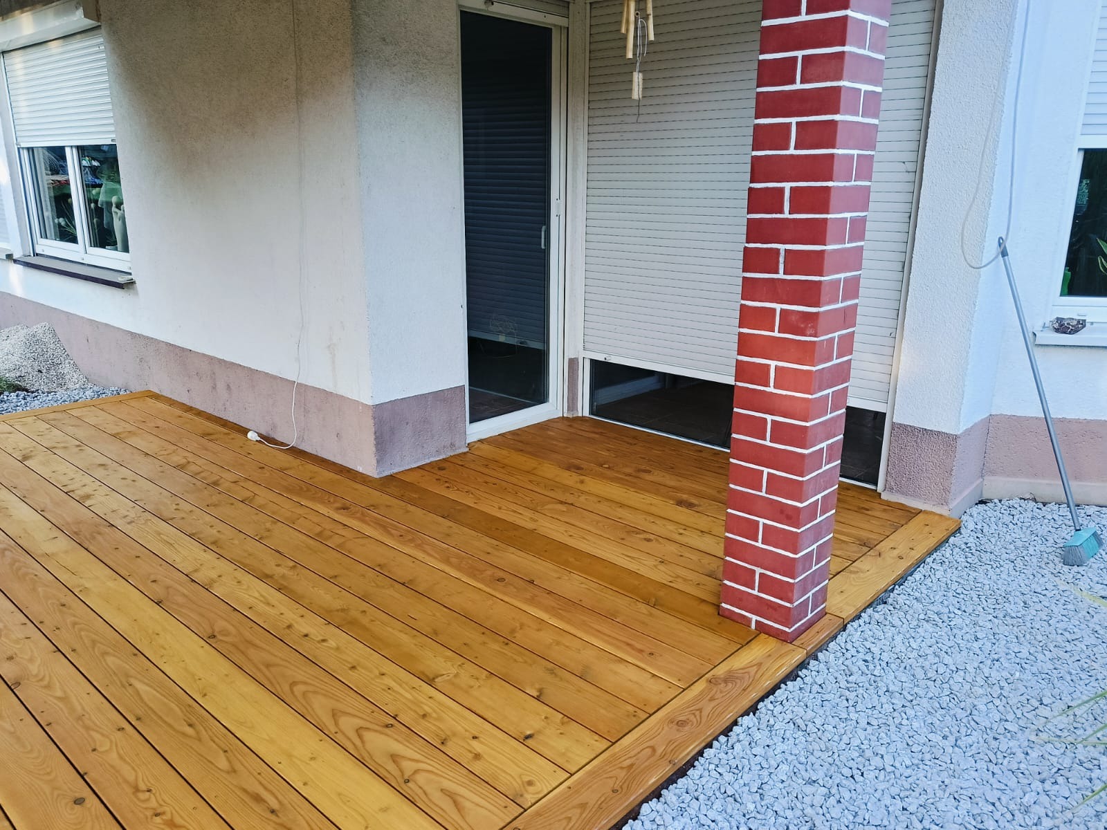 Nowa, drewniana powierzchnia przylegająca do budynku z ceglanym filarem, otoczona drobnym żwirem.