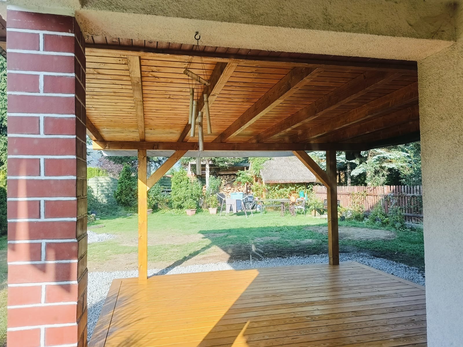 Zadaszony taras z drewnianą podłogą i widokiem na zadbany ogród z elementami małej architektury, w tle widoczny domek z trzcinowym dachem.