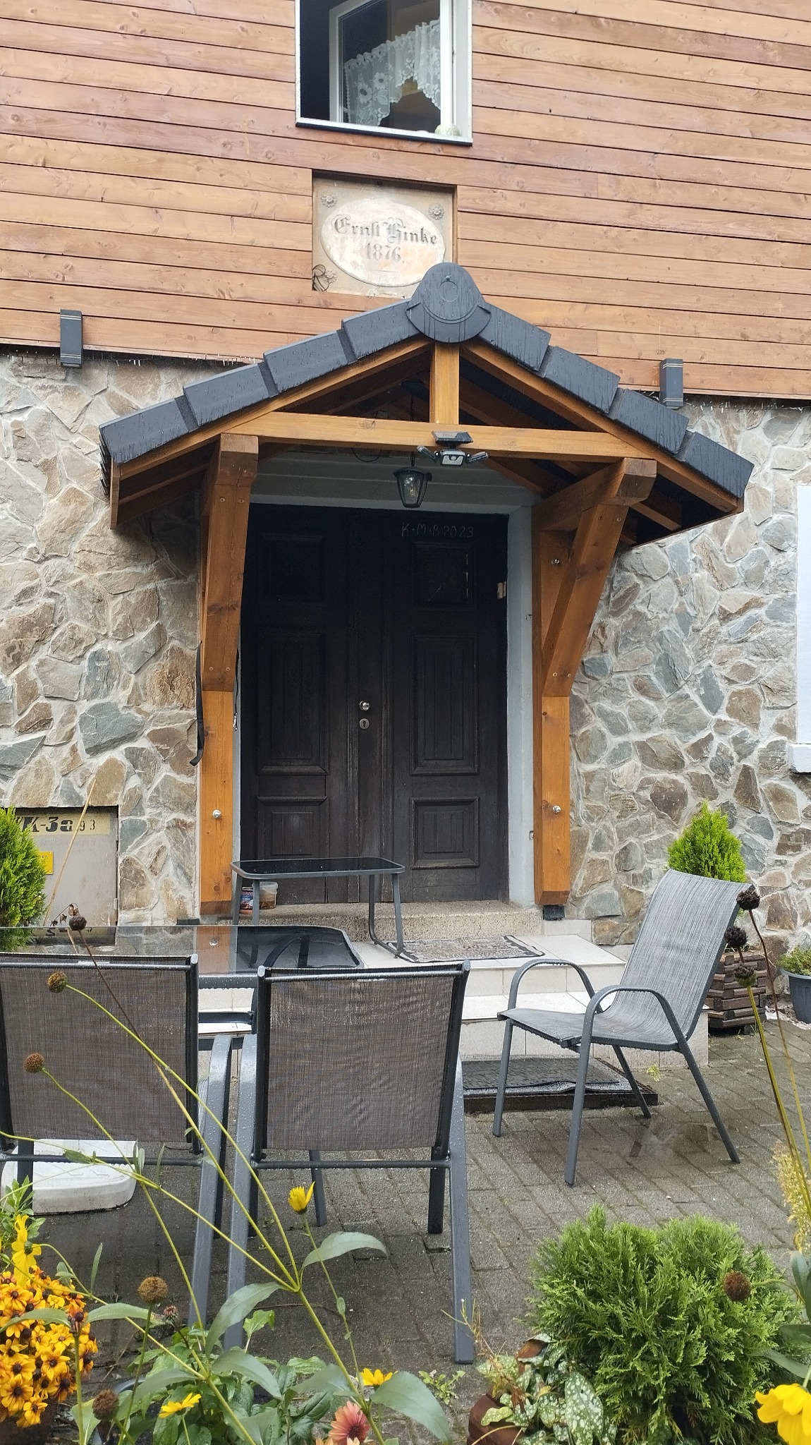 Wejście do domu z kamienną elewacją i drewnianą konstrukcją zadaszenia nad drzwiami, widoczne krzesła ogrodowe i stolik na zewnątrz.