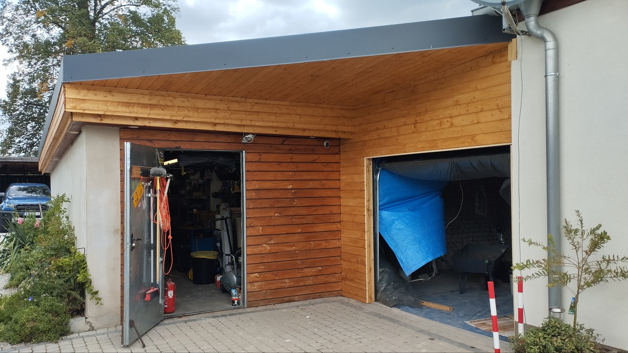 Wykończony garaż z drewnianą elewacją i podbitką, otwarte metalowe drzwi ukazują wnętrze warsztatu, a drugie wejście zasłonięte jest niebieską plandeką.