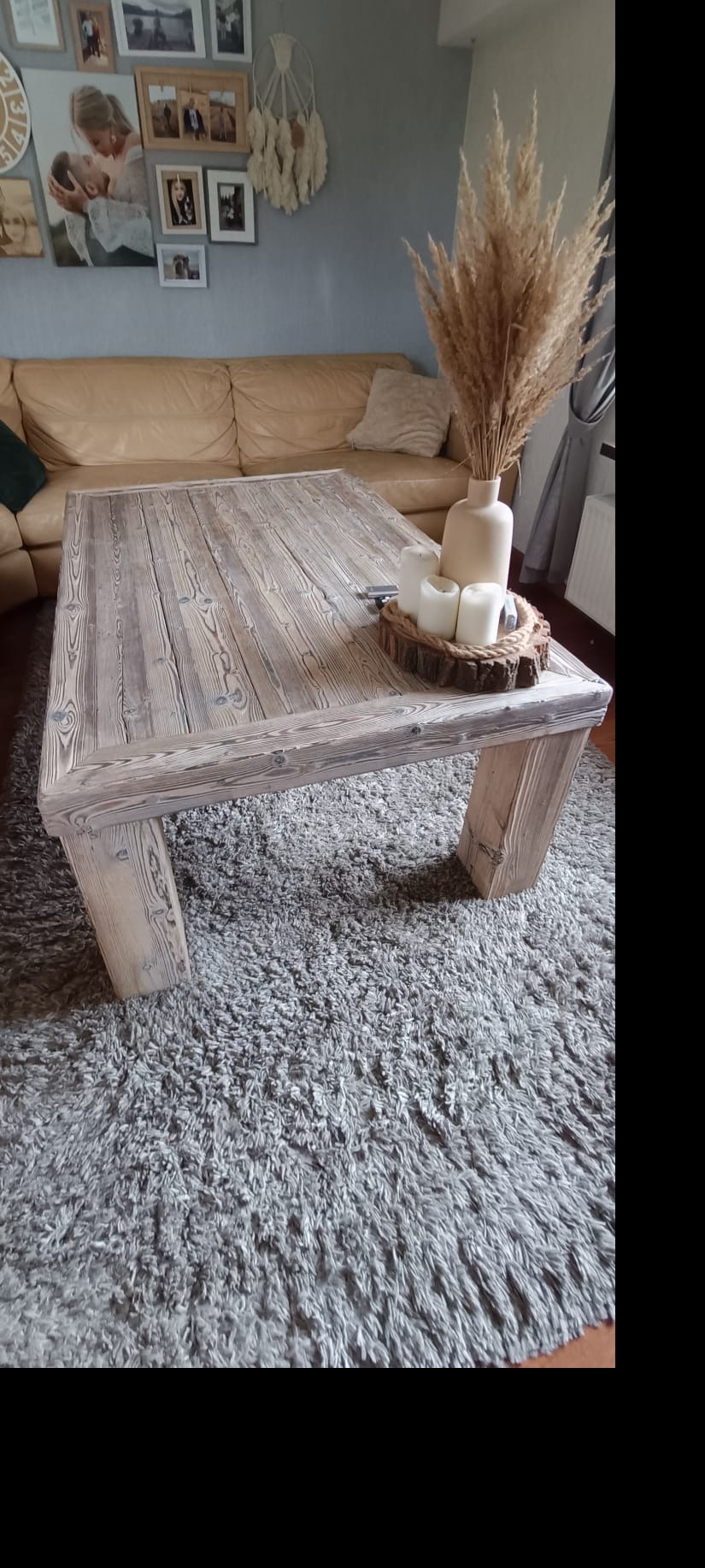 Stolik kawowy z surowego, bielonego drewna, na grubych nogach, ustawiony na puszystym, szarym dywanie. Na blacie dekoracja z suszonych traw w wazonie i świec na drewnianej podstawce. W tle beżowa...