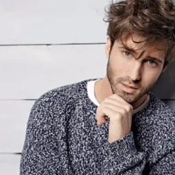 Portret młodego mężczyzny z brązowymi, potarganymi włosami i lekkim zarostem, ubranego w ciemny sweter z białymi plamkami, opierającego brodę na dłoni na tle białej, drewnianej ściany.