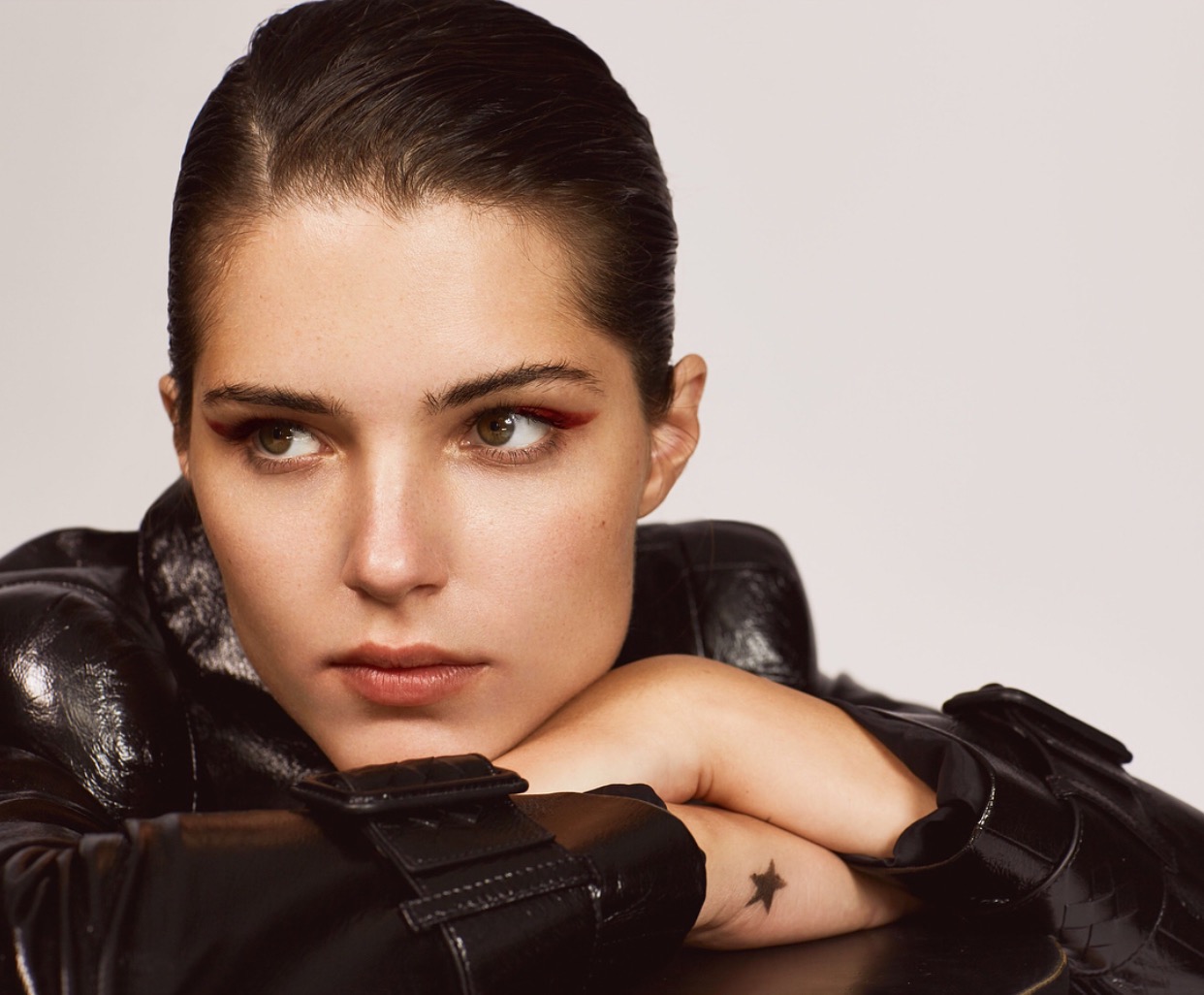 Portret kobiety z gładko zaczesanymi włosami i wyrazistym, czerwonym makijażem oczu, ubrana w lakierowaną, czarną kurtkę.