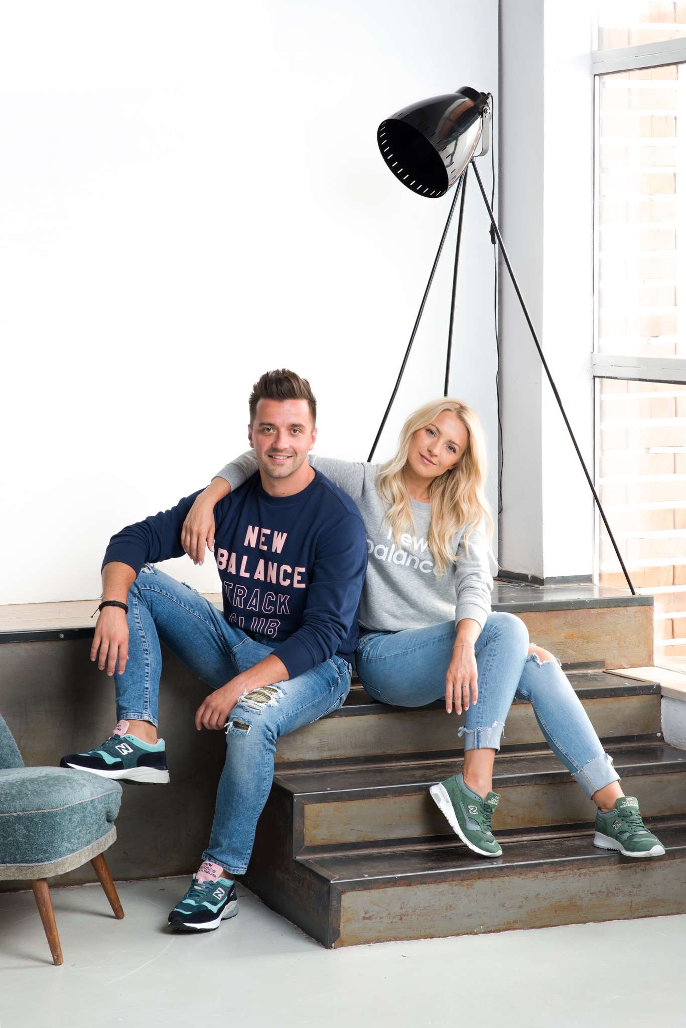 Para siedząca na schodach w dopasowanych bluzach i jeansach New Balance, on w granatowej, ona w szarej, oboje w sportowych butach New Balance w odcieniach zieleni, w tle lampa na trójnogu i okno.