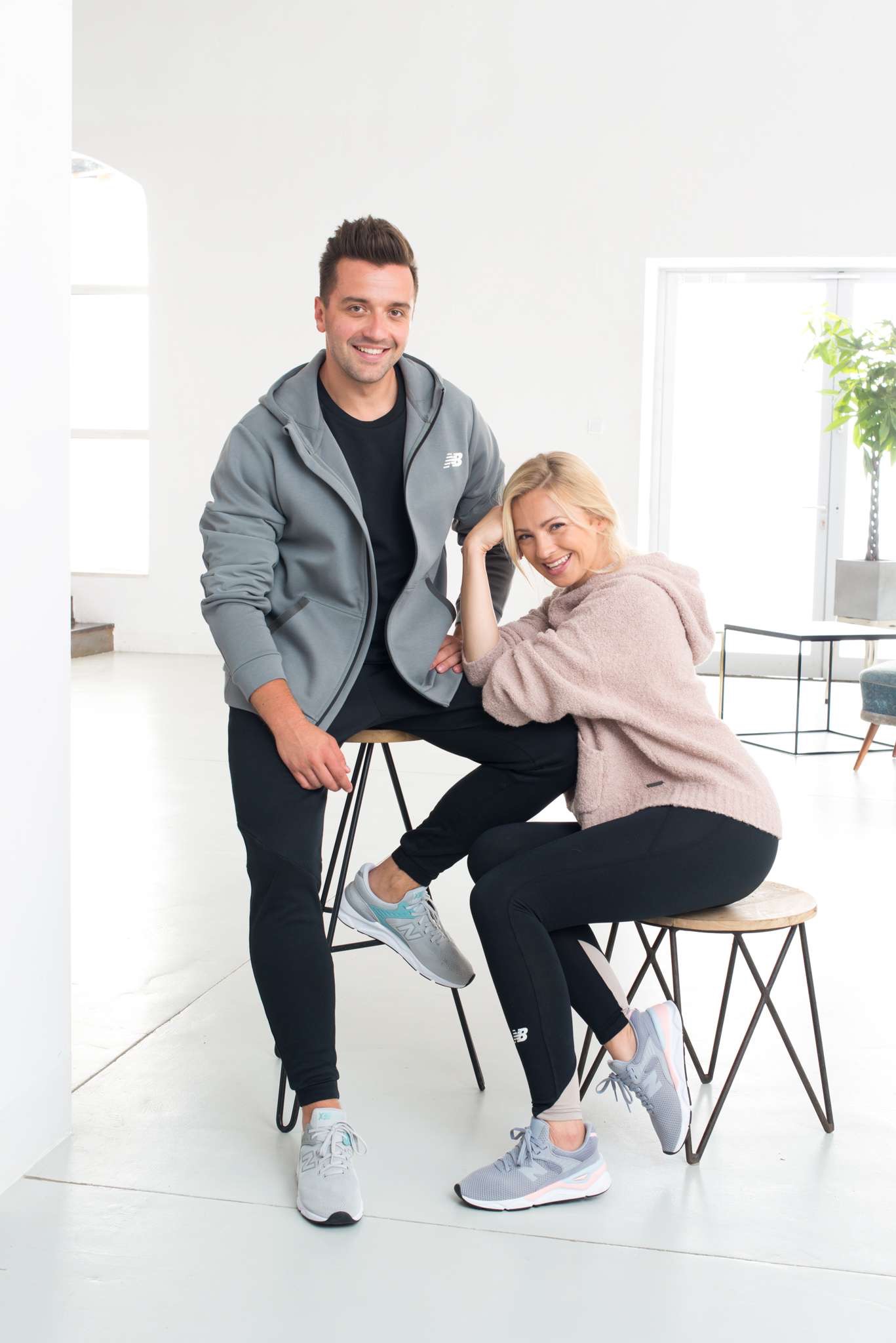 Para w sportowych strojach New Balance pozuje w jasnym wnętrzu, on w szarej bluzie z kapturem i czarnych spodniach, ona w różowej bluzie z kapturem i czarnych legginsach, oboje siedzą na stołkach.