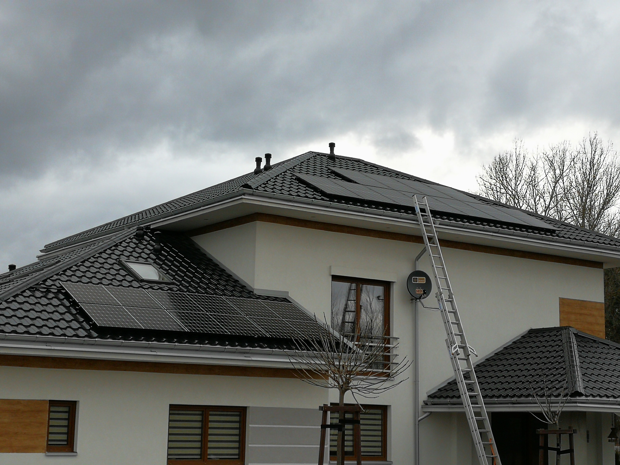 Dom jednorodzinny z zainstalowanymi panelami słonecznymi na dachu pokrytym czarną dachówką, drabina oparta o dach, pochmurne niebo w tle.