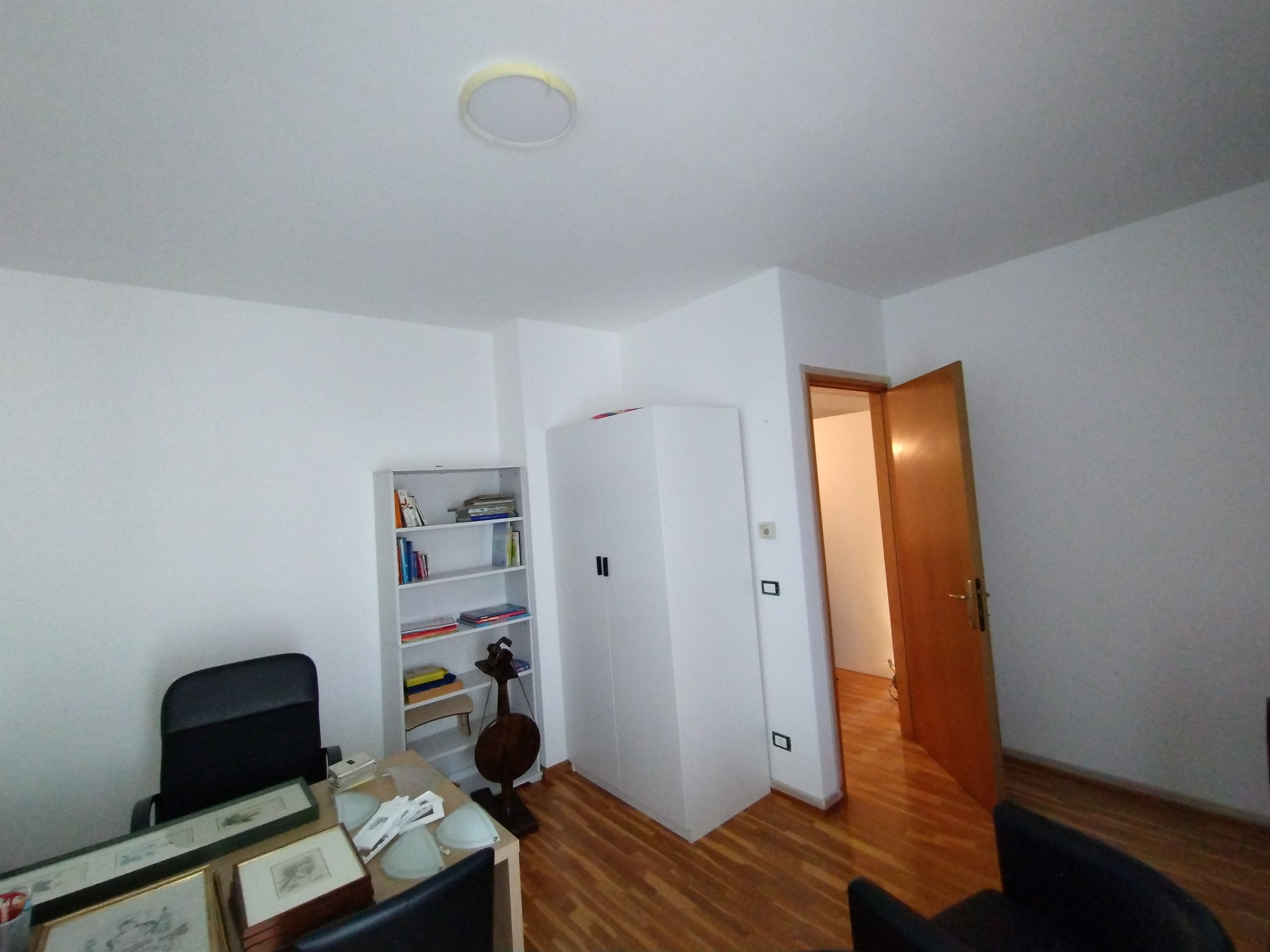 Wnętrze biura z białymi ścianami, drewnianą podłogą, biurkiem z czarnym krzesłem, regałem z książkami i szafą, widok na otwarte drzwi do innego pomieszczenia.
