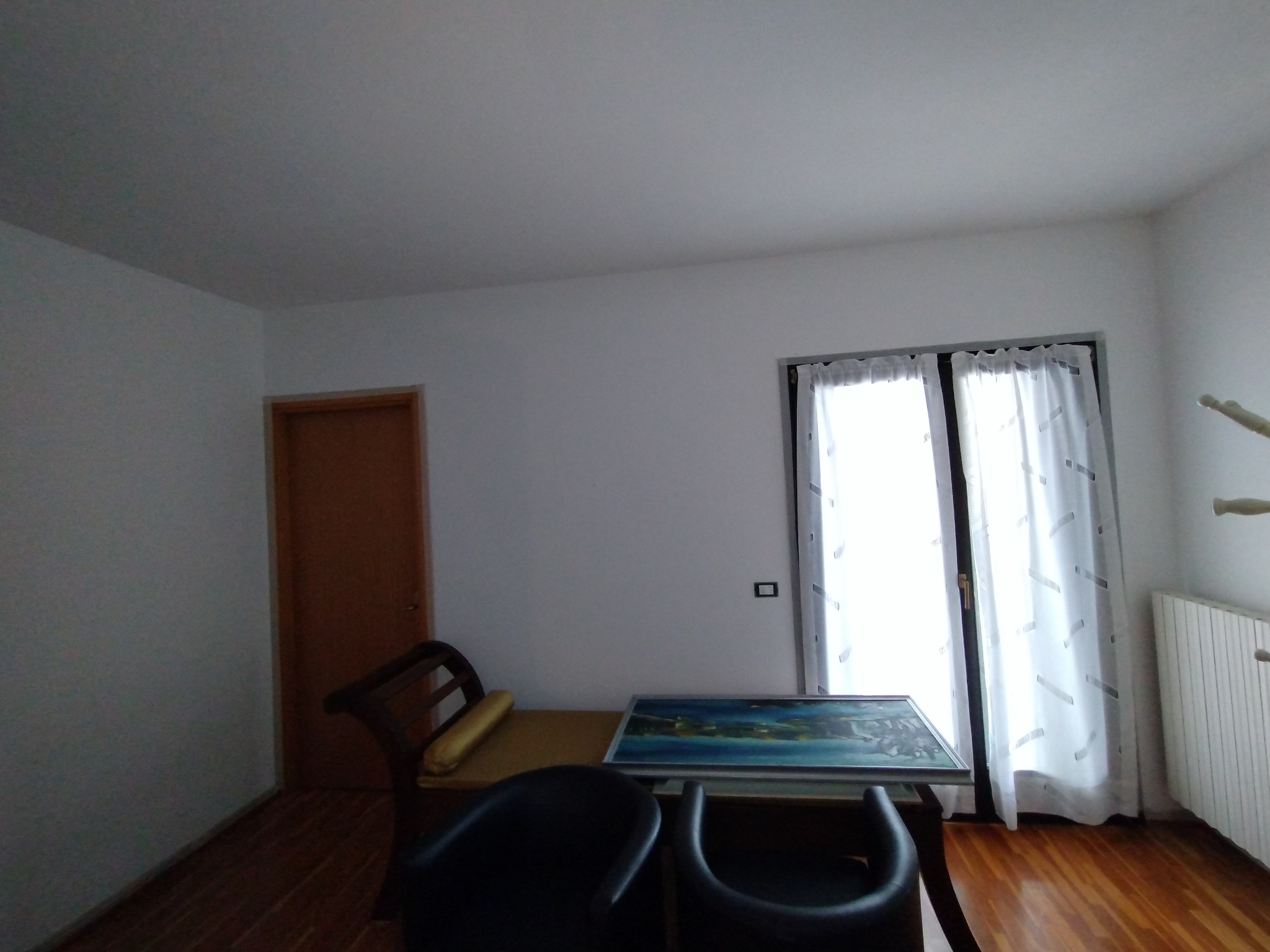 Pokój z białymi ścianami, brązowymi drzwiami i obrazem na stole, oświetlony przez okno z białymi zasłonami.