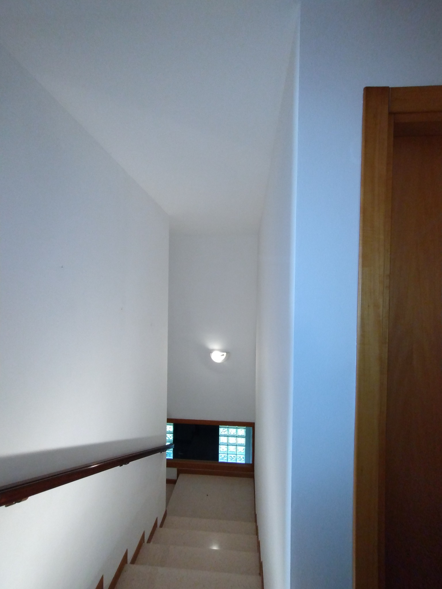Wnętrze klatki schodowej z pomalowanymi na biało ścianami, drewnianą balustradą i oknem z luksferami na końcu schodów.