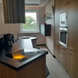 Nowoczesna kuchnia z drewnianymi frontami, czarnym blatem i wbudowanym sprzętem AGD, widok na okno z zielenią za nim.