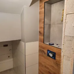 Wnętrze łazienki w trakcie prac wykończeniowych: widoczna zabudowa z drewnopodobnych paneli z wnęką, częściowo wykończone płytki na ścianach, surowy mur z boku, instalacja sanitarna w trakcie montażu.