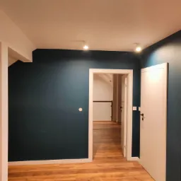 Pomieszczenie po odświeżeniu: ściany pomalowane na ciemny granat, jasne panele podłogowe, białe drzwi i ościeżnice, widoczne elementy wykończeniowe.