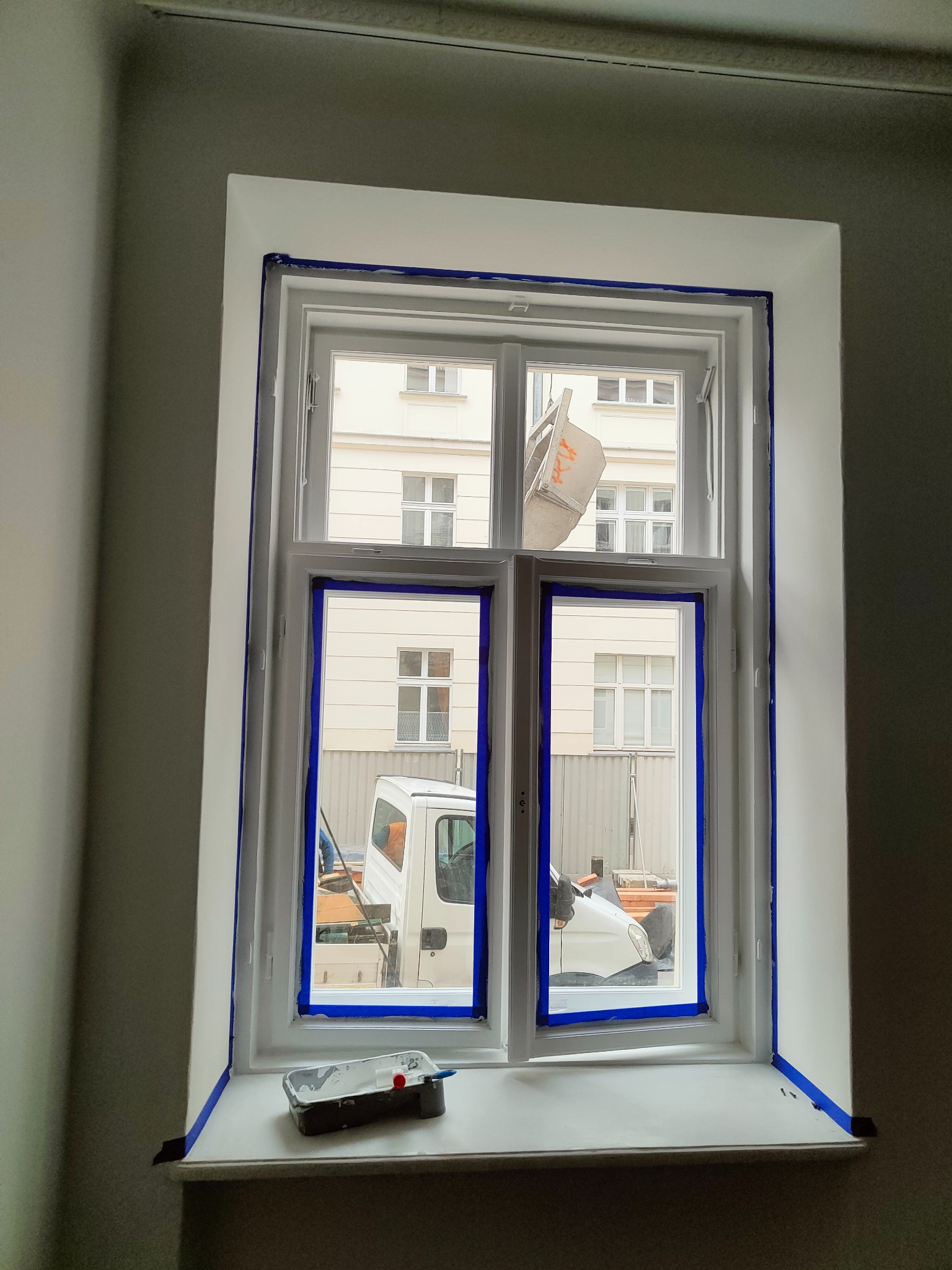 Przygotowane do malowania okno z niebieską taśmą malarską, na parapecie kuweta z farbą i wałkiem, widok na budynek i zaparkowany samochód dostawczy za oknem.