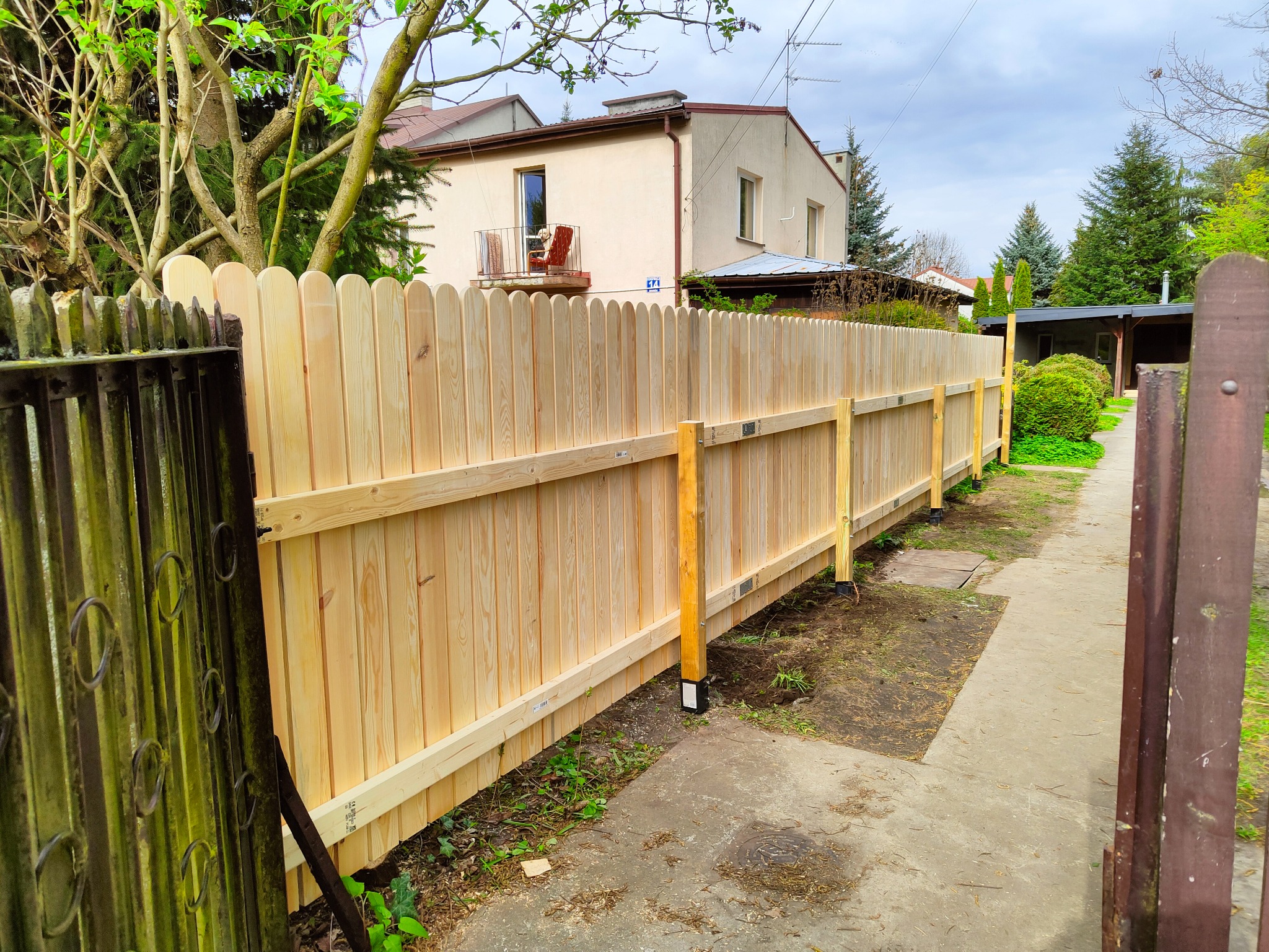 Nowo postawiony, surowy, drewniany płot lamelowy, widok z perspektywy furtki z metalowymi, rdzewiejącymi prętami, w tle dom z balkonem.