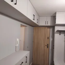 Przedpokój z szarymi ścianami, zabudową szafek pod sufitem z czarnymi uchwytami, drewnianymi drzwiami wejściowymi i szafką na buty z wieszakami.