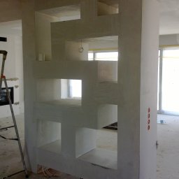 Eko-Bud Energy - Surowa ściana działowa z geometrycznymi otworami, widoczne przewody elektryczne, drabina w tle, stan deweloperski.