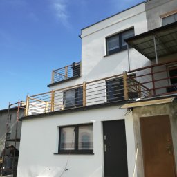 Eko-Bud Energy - Dwupiętrowy dom z białymi ścianami i drewnianymi balustradami balkonów, widoczni pracownicy na rusztowaniu po lewej stronie, sugerujący trwające prace wykończeniowe.