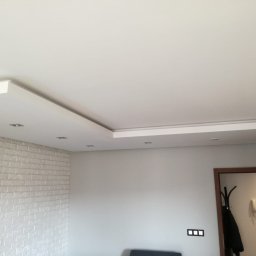 Eko-Bud Energy - Nowoczesny sufit podwieszany z oświetleniem punktowym, ściana z białej cegły dekoracyjnej, widok na fragment pokoju z drzwiami i wieszakiem.