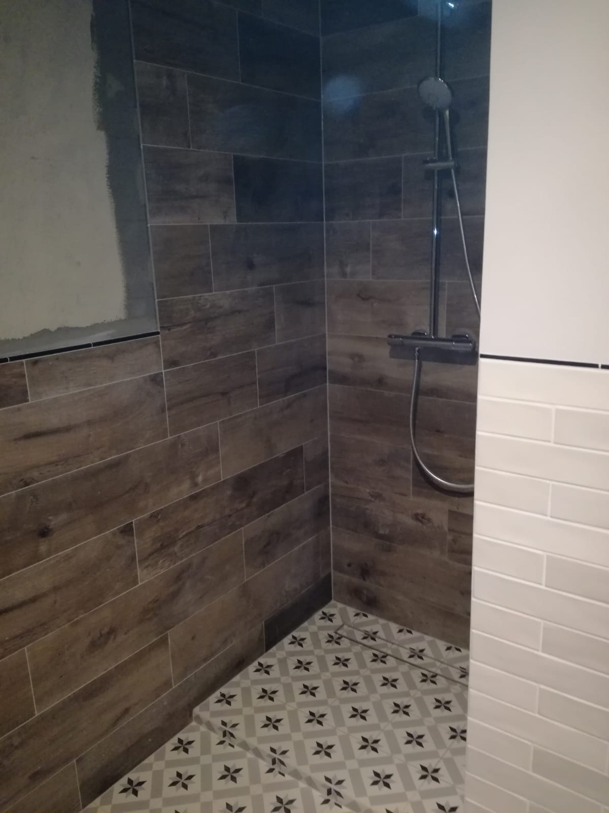 Nowoczesna kabina prysznicowa z brązowymi płytkami imitującymi drewno na ścianach, kontrastującymi białymi płytkami na jednej ze ścian i wzorzystą podłogą z odpływem liniowym.