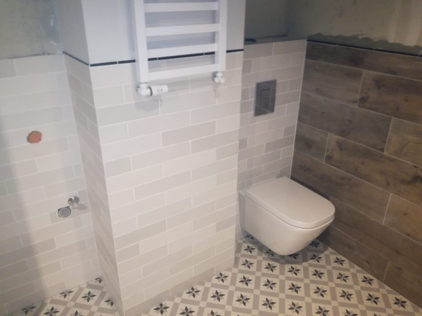 Wykończona łazienka z białą wiszącą toaletą, grzejnikiem drabinkowym, jasnymi płytkami ściennymi ułożonymi w cegiełkę i dekoracyjną podłogą w gwiazdki.