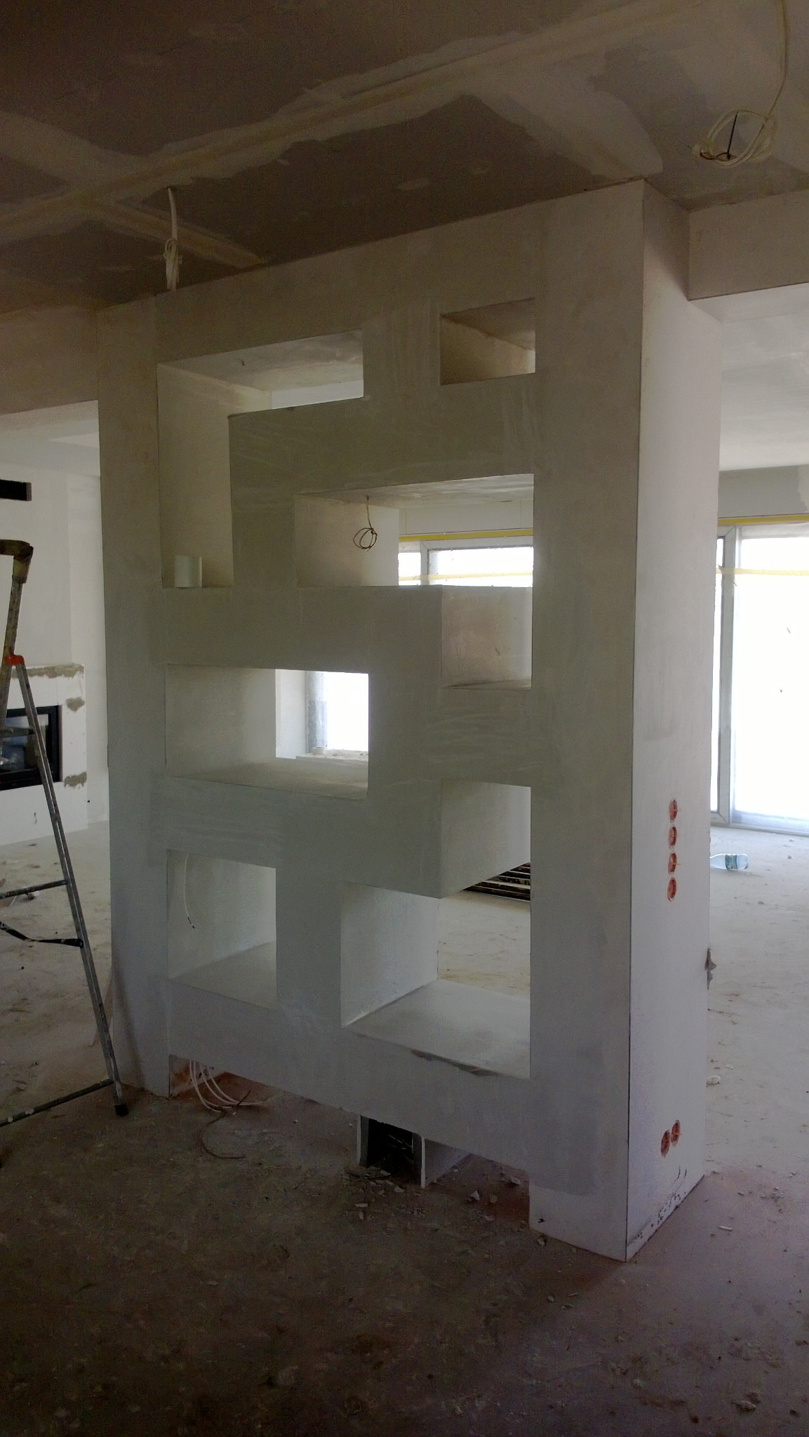 Surowa ściana działowa z geometrycznymi otworami, widoczne przewody elektryczne, drabina w tle, stan deweloperski.