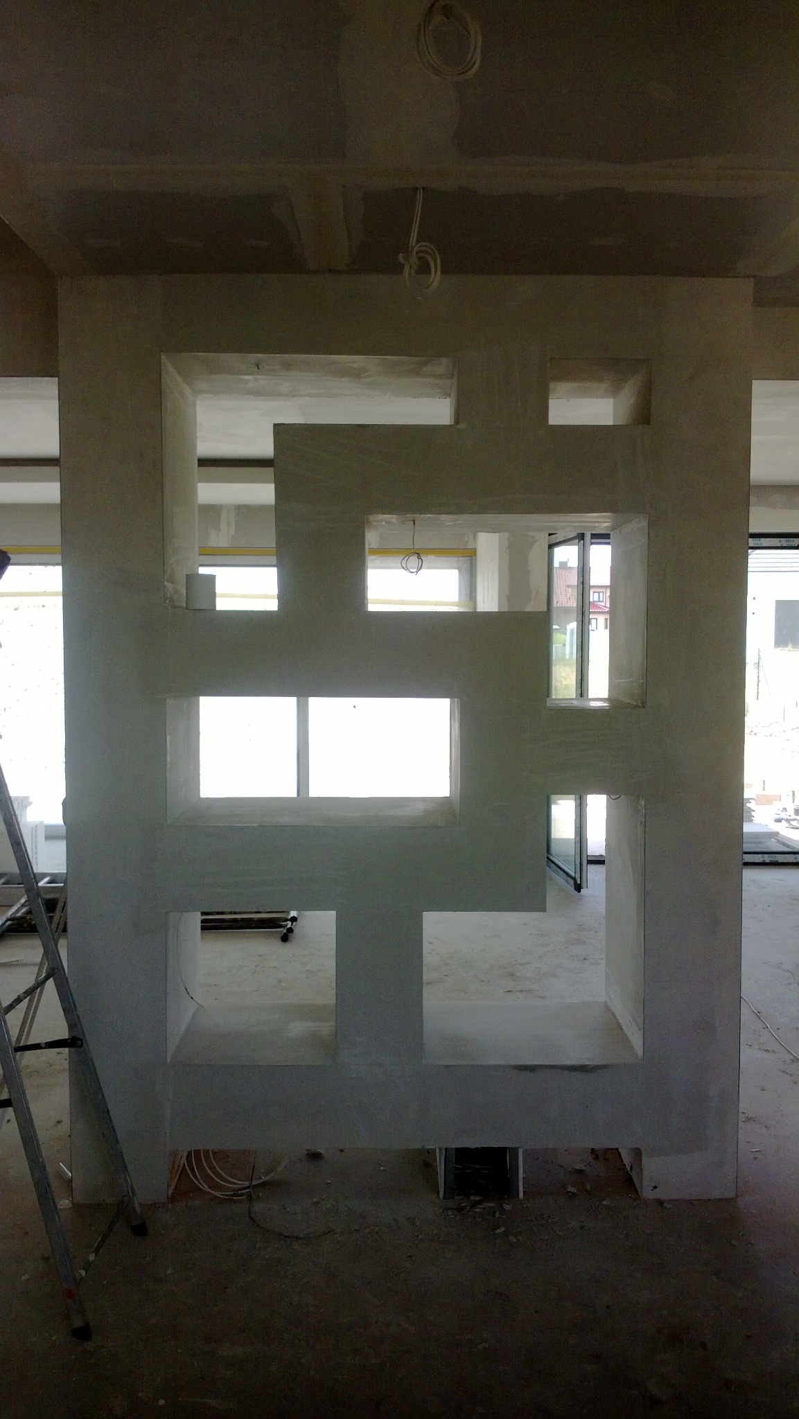 Surowa ściana działowa z geometrycznymi otworami w stanie deweloperskim, widoczne przewody elektryczne i drabina aluminiowa.