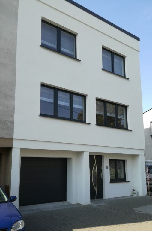 Dwupiętrowy dom z płaskim dachem i garażem, wykończony białą elewacją, z ciemnymi oknami i drzwiami, widok z zewnątrz.